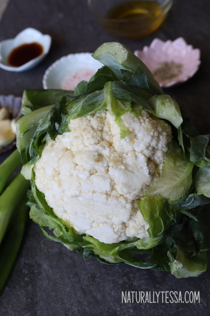 Slate background with the ingredients for garlic cauliflower fried rice: a head of cauliflower, a white bowl of pink Himalayan salt, a pink bowl of black pepper, a glass bowl with olive oil, and fresh scallions.