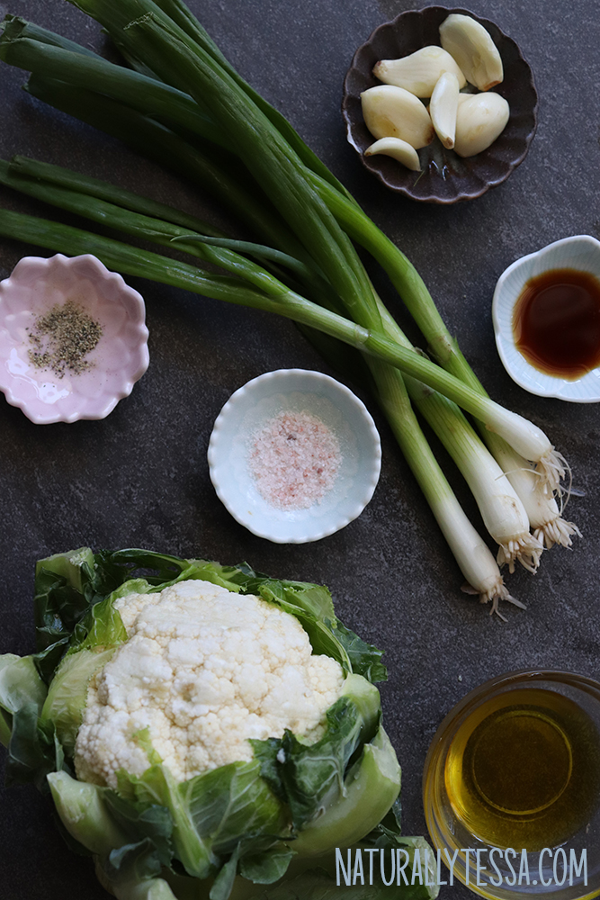 Slate background with the ingredients for garlic cauliflower fried rice: a head of cauliflower, a white bowl of pink Himalayan salt, a pink bowl of black pepper, a glass bowl with olive oil, and fresh scallions.
